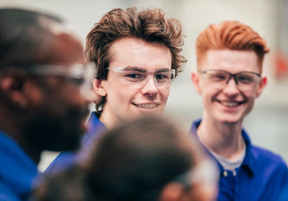 A young man, who is wearing protective eyewear, smiles as he enjoys his engineering class with peers - they are all wearing blue coveralls.