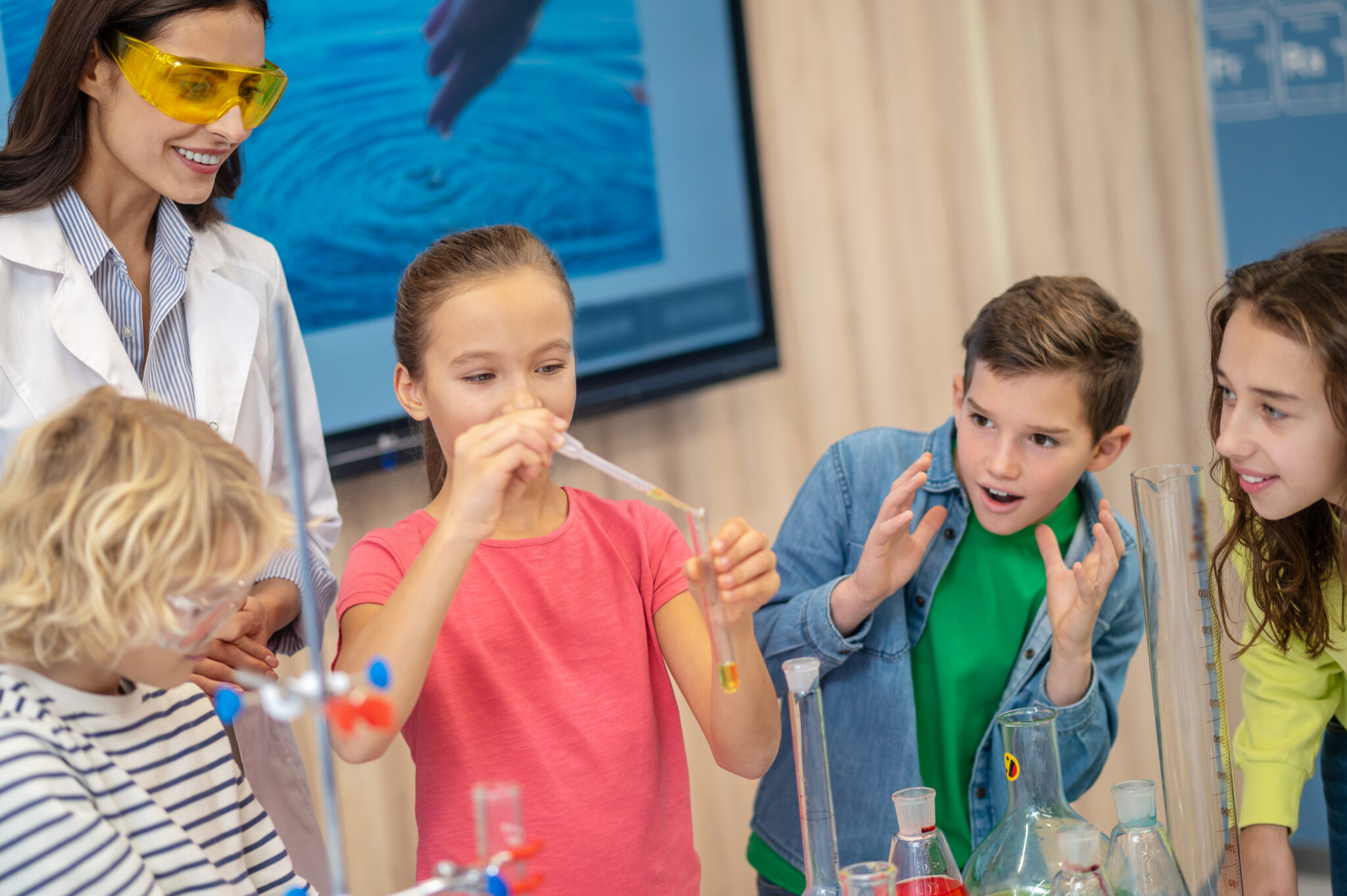 Interesting here. Girl with pipette and test tube and admiring watching children with teacher standing near table in classroom